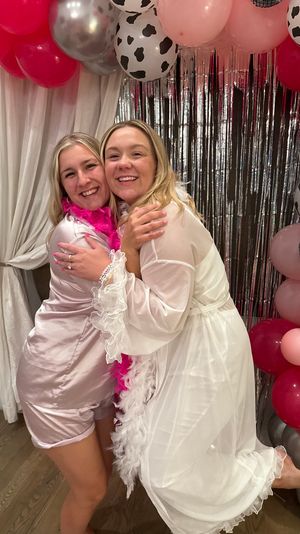 Two smiling women hugging at an indoor party, wearing satin pajamas and a sheer white robe with feather boas, posed in front of a silver fringe backdrop and a pink, silver and cow-print balloon arch.