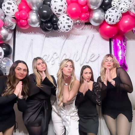 Five friends pose at a bachelorette party photo booth under a pink, black and silver balloon garland; bride-to-be in a white sash blows a kiss while friends in black dresses strike playful prayer-hand poses against a geometric backdrop.