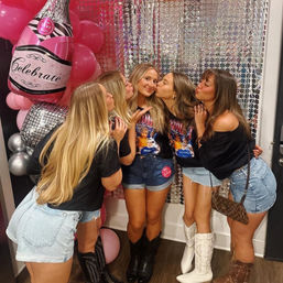 Five friends in denim shorts and cowboy boots pose at a birthday photo booth with pink "Celebrate" balloons and a sparkly sequin backdrop