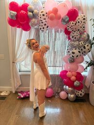Person in white mini dress and glitter cowboy boots showing a ring in front of a pink, silver and cow-print balloon arch with metallic fringe indoor party backdrop