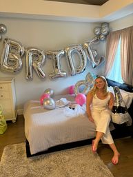 Home bedroom bachelorette scene with large silver "BRIDE" balloons over the bed, pink and silver balloons, bridal hat and veil, and a smiling woman in white holding an inflatable champagne bottle by the window.