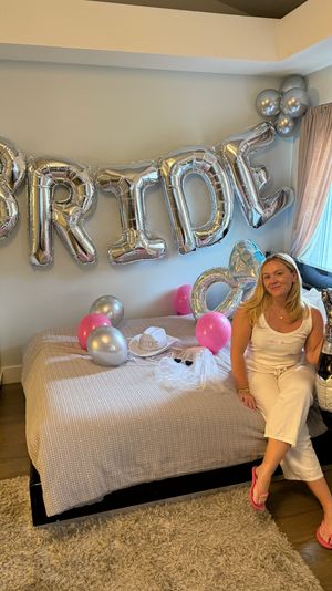 Home bedroom bachelorette scene with large silver "BRIDE" balloons over the bed, pink and silver balloons, bridal hat and veil, and a smiling woman in white holding an inflatable champagne bottle by the window.