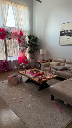 Sunlit modern living room with pink, silver, and patterned balloon arch by a tall window; leather sectional, wooden coffee table strewn with pink party decorations, gift bags, cowboy boot balloons, and feathers on the rug — post‑celebration scene.