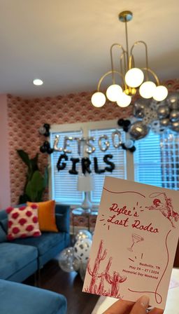 Handheld pink ‘Last Rodeo’ party invitation for a Nashville Memorial Day weekend celebration; festive living room in background with “LET'S GO GIRLS” balloons, silver and black balloons, blue velvet sofa with colorful pillows, patterned pink wallpaper and a modern brass chandelier.
