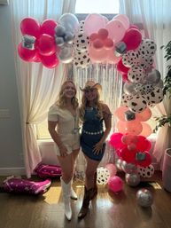 Two women in cowgirl outfits (white romper with white boots, denim dress with cowboy hat and boots) smiling under a pink, silver and cow‑print balloon arch with metallic fringe photo backdrop at an indoor party.
