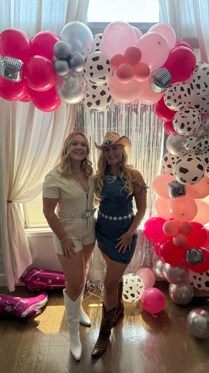 Two women in cowgirl outfits (white romper with white boots, denim dress with cowboy hat and boots) smiling under a pink, silver and cow‑print balloon arch with metallic fringe photo backdrop at an indoor party.