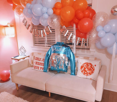 Cheerful living room bachelorette setup with orange and pale blue balloon garland above a cream sofa, metallic 'Team Bride' jacket balloon on cushions, decorative pillows, pink shag rug and wood floor.