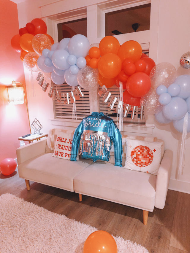 Cheerful living room bachelorette setup with orange and pale blue balloon garland above a cream sofa, metallic 'Team Bride' jacket balloon on cushions, decorative pillows, pink shag rug and wood floor.