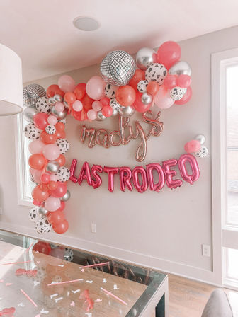 Playful pink and silver balloon garland with mirrored disco balls and letter balloons above a confetti-sprinkled glass dining table in a bright modern home — indoor party decoration.