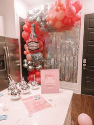 Indoor pink and silver birthday party setup in a home kitchen with a balloon arch, champagne-shaped foil balloon, silver sequin backdrop, disco-ball drink cups and a pink event sign on the counter.
