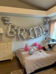 Cozy bedroom decked out for a bachelorette: giant silver foil 'BRIDE' letters on the wall, pink and silver balloons, inflatable diamond ring and champagne bottle, bridal hat and veil on the bed.