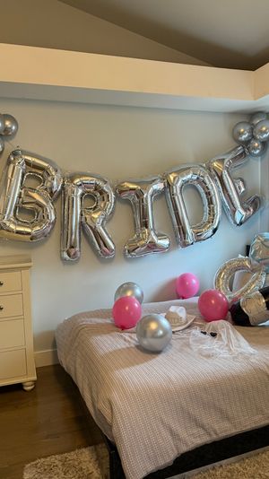 Cozy bedroom decked out for a bachelorette: giant silver foil 'BRIDE' letters on the wall, pink and silver balloons, inflatable diamond ring and champagne bottle, bridal hat and veil on the bed.