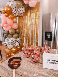 Festive Nashville party setup in a kitchen: pink, gold and cow-print balloon garland, gold fringe backdrop, rose-gold disco ball drink cups on a marble island, and a smiling face paddle next to a welcome sign.