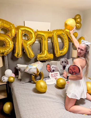 Bachelorette party bedroom: bride-to-be kneeling on bed amid large gold "BRIDE" balloons, balloon ring, scattered gold balloons and playful face-cutout props.