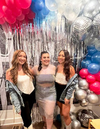 Three women smiling at an indoor party in front of a silver tinsel fringe backdrop and colorful balloon arch with disco-ball balloons, wearing sparkly outfits and denim jackets, celebratory event photo.