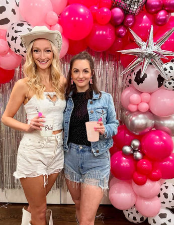 Two smiling women posing at a bachelorette-style party in front of a pink, silver and cow-print balloon arch and metallic fringe backdrop; left in a white cowgirl hat and white outfit holding a cup labeled 'Bride', right in a denim jacket and beaded fringe shorts holding a pink cup.