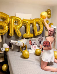 Playful bachelorette bedroom scene: woman in a white dress kneeling on a bed surrounded by large gold "BRIDE" foil balloons, gold and white balloons, a ring-shaped balloon and face and dog cutouts.