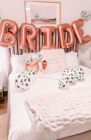 Bedroom decorated for a bachelorette party with rose-gold BRIDE balloons, cow-print balloons, a diamond-ring foil balloon, faux-fur throw and a pillow reading "Future Mrs." on a white bed.