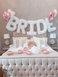 Tufted bridal suite bed styled for wedding morning with oversized white 'BRIDE' letter balloons above, rose-gold heart and white balloons on and around the bed.