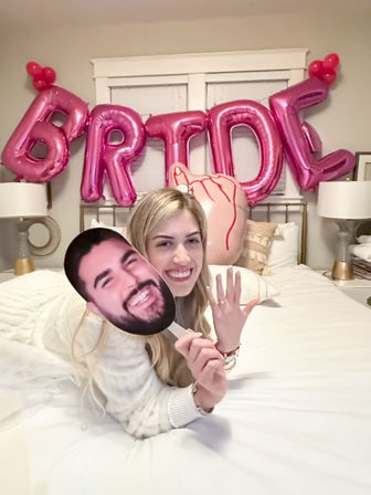 Smiling woman lying on a bed in a cozy bedroom, holding a large face cutout and showing an engagement ring in front of metallic pink “BRIDE” balloons and a heart-shaped balloon.