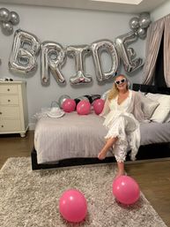 Bride-to-be in a white robe and heart-shaped sunglasses sits on a bed in a decorated bedroom with large silver 'BRIDE' foil balloons and scattered pink balloons for a bachelorette celebration.