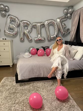 Bride-to-be in a white robe and heart-shaped sunglasses sits on a bed in a decorated bedroom with large silver 'BRIDE' foil balloons and scattered pink balloons for a bachelorette celebration.