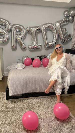 Bride-to-be in a white robe and heart-shaped sunglasses sits on a bed in a decorated bedroom with large silver 'BRIDE' foil balloons and scattered pink balloons for a bachelorette celebration.