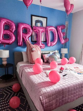 Bedroom party setup with metallic pink letter balloons on a blue wall, pink round balloons scattered across a pink-quilted bed with a disco-ball pillow and bedside lamps.