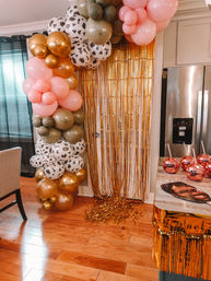 Festive indoor kitchen party scene with pink, gold, olive and cow-print balloon garland and shimmering gold fringe doorway backdrop over hardwood floors.