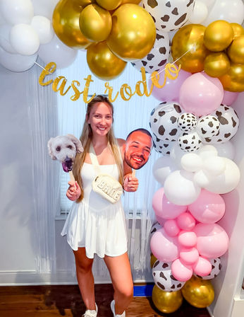 Smiling woman in a white dress wearing a 'BRIDE' crossbody bag, holding a dog head and a smiling face photo prop beneath a gold "last rodeo" banner and pink, white, cow-print and gold balloon arch.