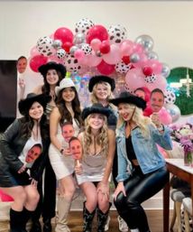 Group shot of women at an indoor cowgirl-themed celebration under a pink, red and cow-print balloon arch, wearing cowboy hats and boots and holding face-cutout photo booth props.