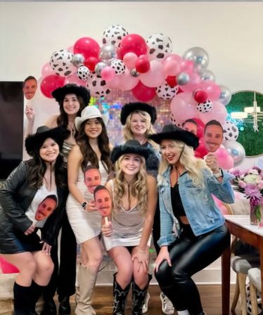 Group shot of women at an indoor cowgirl-themed celebration under a pink, red and cow-print balloon arch, wearing cowboy hats and boots and holding face-cutout photo booth props.