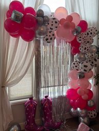 Indoor party backdrop with a pink and silver balloon arch featuring cow‑print balloons, mirrored disco ball accents and a silver fringe curtain over a window, plus inflatable pink cowboy boots on the floor.