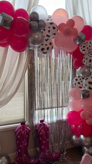 Indoor party backdrop with a pink and silver balloon arch featuring cow‑print balloons, mirrored disco ball accents and a silver fringe curtain over a window, plus inflatable pink cowboy boots on the floor.