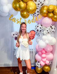 Smiling woman in a white dress at an indoor bachelorette party photo spot with a gold, pink, white and cow-print balloon arch, glitter “last rodeo” banner and dog and face cutout props