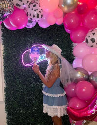 Bachelorette party photo: woman in a denim lace dress and white veiled cowboy hat kisses a face cutout in front of a green foliage wall, pink and cow-print balloon arch, neon sign and disco-ball detail.