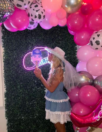 Bachelorette party photo: woman in a denim lace dress and white veiled cowboy hat kisses a face cutout in front of a green foliage wall, pink and cow-print balloon arch, neon sign and disco-ball detail.