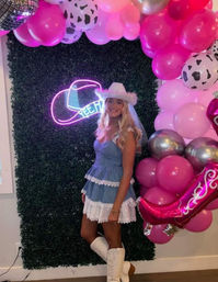 Woman in denim lace mini dress, white cowgirl hat and boots posing at an indoor party photo wall with a neon cowboy hat sign and pink, silver and cow-print balloons.