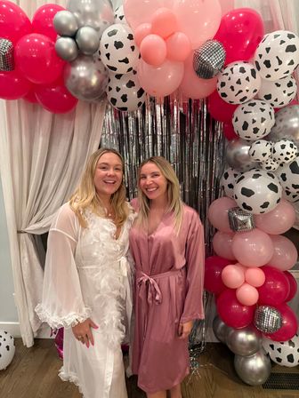 Two smiling friends in a white feather-trimmed robe and pink satin robe pose by a metallic fringe backdrop and a festive balloon arch of pink, silver and cow-print balloons at an indoor party.