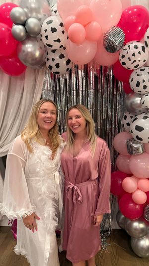 Two smiling friends in a white feather-trimmed robe and pink satin robe pose by a metallic fringe backdrop and a festive balloon arch of pink, silver and cow-print balloons at an indoor party.