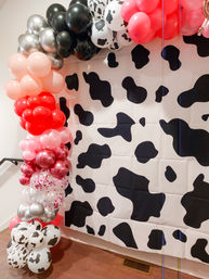 Playful indoor cow-print photo backdrop framed by a bubbly pink, red, silver, black and cow-spotted balloon garland arch for party decor.