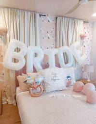 Bridal-themed bedroom in pastel pink with large white 'BRIDE' letter balloons over the bed, rose-gold ring-shaped balloon, pink round balloons, patterned pillows, soft curtains and floral wallpaper.