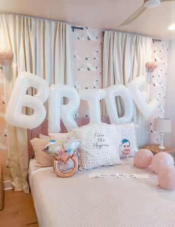 Bridal-themed bedroom in pastel pink with large white 'BRIDE' letter balloons over the bed, rose-gold ring-shaped balloon, pink round balloons, patterned pillows, soft curtains and floral wallpaper.