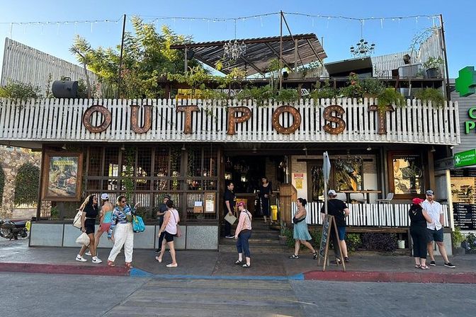 Two-story coastal eatery with white picket facade, oversized marquee letters, rooftop garden and string lights; pedestrians and diners gather on the sunny sidewalk in a lively beachside shopping strip.