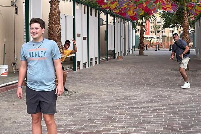 Three friends posing and joking in a palm-lined downtown pedestrian alley with brick paving and a colorful umbrella canopy overhead.
