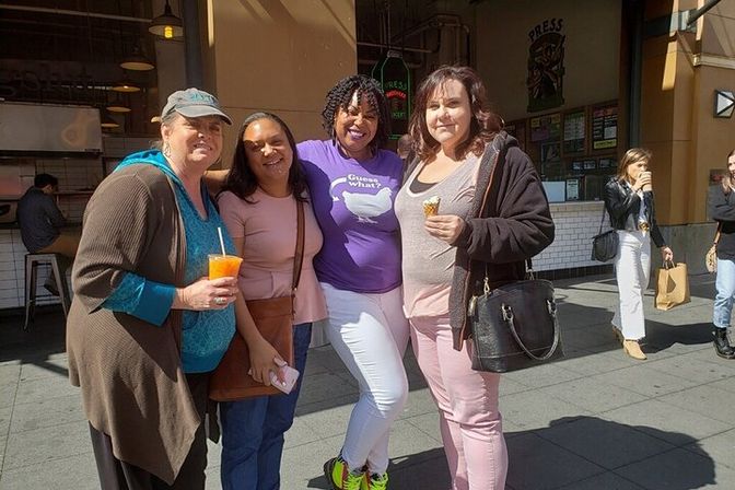 Four women smiling together outside a busy urban food market on a sunny day, two holding snacks — a bright orange drink and an ice cream cone — casual outfits with shoppers in the background.