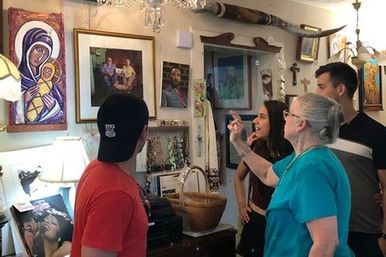 Four people browse and point at eclectic wall art and religious icons inside a cozy antique shop, surrounded by framed paintings, a jewelry display, wicker baskets, a chandelier and longhorn decor.