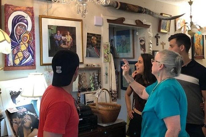 Four people browse and point at eclectic wall art and religious icons inside a cozy antique shop, surrounded by framed paintings, a jewelry display, wicker baskets, a chandelier and longhorn decor.