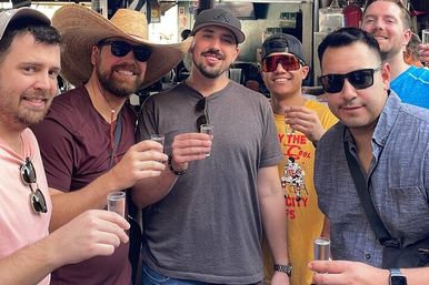 Group of six men smiling and raising shot glasses at a lively outdoor bar, casually dressed with sunglasses and a large straw cowboy hat