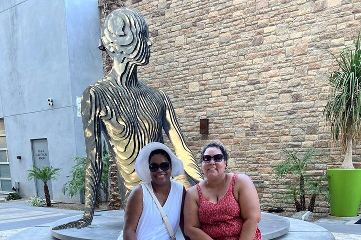 Two smiling women in sunglasses, one in a white sunhat, seated in front of a large layered metal human sculpture in an outdoor urban courtyard with a stone wall and potted palms.